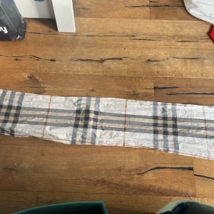 Burberry  - Lång och tunn sjal i beige med rutigt mönster i svart, vitt och brunt. Tillverkad i ett lätt och mjukt material som passar bra att ha runt halsen eller över axlarna. Perfekt accessoar för att lyfta en outfit.