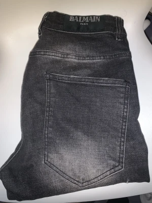 Svarta jeans från Balmain 29/32 - Säljer ett par svarta slim fit jeans från Balmain med snygga slitningar och en cool dragkedjedetalj på framsidan. Jeansen har klassisk femficksdesign och är tillverkade i mjukt jeansmaterial. Perfekta för dig som vill ha en edgy och trendig look.
