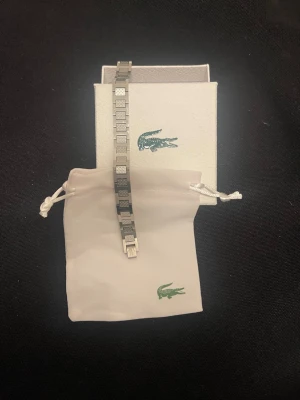 Silverfärgat armband från Lacoste - Snyggt armband i silverfärgad metall från Lacoste med rektangulära länkar och diskret krokodillogga. Kommer med original ask och tygpåse med Lacoste-logga. Perfekt accessoar för en stilren look. Använd några fåtal gånger och säljer pågrund av att jag fått en ny klocka som gör att jag inte har någon användning för det.