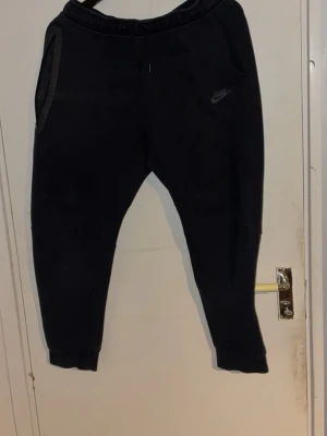 Svarta mjukisbyxor från Nike - Svarta mjukisbyxor från Nike med diskret logga på vänster ben. Byxorna har resår i midjan, snörning och muddar vid bensluten. Tillverkade i mjukt material som passar perfekt för chill eller träning. Två fickor med dragkedja för säker förvaring.