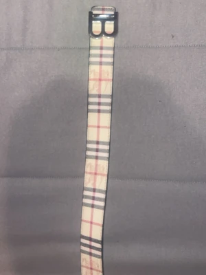 Rutmönstrat bälte från Burberry - Snyggt bälte från Burberry med klassiskt rutmönster i beige, svart och rött. Bältet har en silverfärgad metallspänne och är tillverkat i skinn. Perfekt accessoar för att lyfta din outfit.