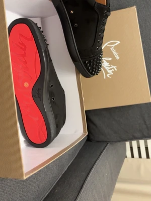 Svarta Louboutin  - Säljer ett par svarta sneakers från Christian Louboutin med klassisk röd sula och svarta nitar på tån. Skorna har snörning och är tillverkade i mocka för en lyxig känsla. Perfekta för dig som vill sticka ut med en edgy och exklusiv look.