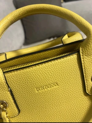 Gul handväska från Don Donna - Jag säljer en fin väska som kostade 499 kr ny. Den är endast använd en gång och är i mycket fint skick. Säljes för 199 kr .Snygg gul handväska från Don Donna i strukturerat skinn. Väskan har både handtag och en axelrem med kedjedetaljer, vilket ger en cool vibe. Perfekt storlek för att få plats med allt du behöver och stilren design med svarta kanter.