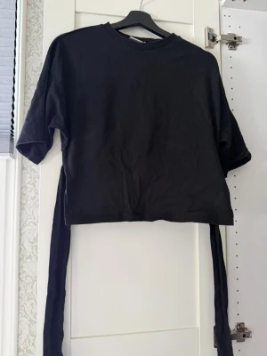Svart croppad topp med knytband - Säljer en svart croppad topp med långa knytband i midjan som kan knytas bak. Toppen har rund halsringning och korta ärmar. Perfekt för dig som gillar en enkel men trendig look.