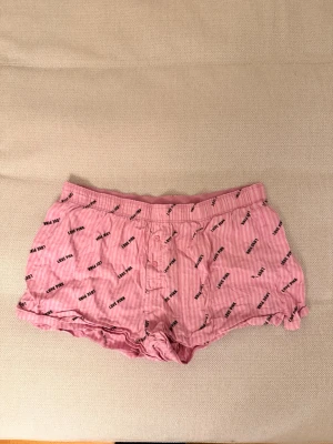 Rosa boxershorts från Victoria’s secret PINK - Säljer ett par rosa boxershorts från Victoria’s secret PINK med tunna vita ränder och svart textmönster där det står 'LOVE PINK' och 'PINK 1986' över hela tyget. De har knappgylf framtill och resår i midjan. Midjemått: 40cm, längd: 25cm 💖