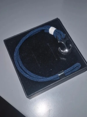 Blått armband med ankardetalj - Snyggt armband med marinblått flätat band och en silverfärgad ankare som lås. Detalj i vitt snöre vid ankaret ger en cool kontrast. Perfekt accessoar för dig som gillar nautisk stil och vill sticka ut lite extra.