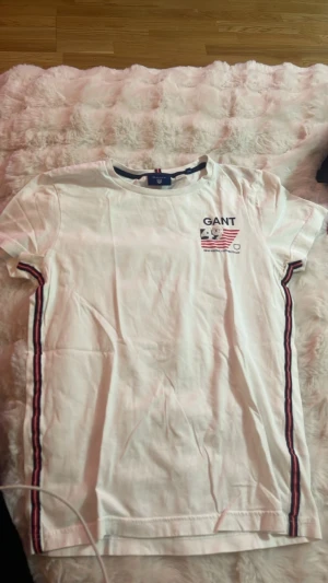 Vit GANT t-shirt med broderi - Vit t-shirt från GANT med broderad logga och siffran 49 på bröstet. Snygga ränder i rött, blått och vitt längs sidorna. Klassisk passform och rund halsringning. Perfekt för en sportig och stilren look.