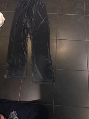 Mörkgrå velour mjukisbyxor - Säljer ett par mörkgrå mjukisbyxor i mjuk och glansig velour. Byxorna har resår i midjan och vida ben för en chill och avslappnad look. Perfekta för att mysa runt hemma eller hänga med vänner.det finns även en likadan tröja med huva och storleken på tröjan/byxorna är i S.
