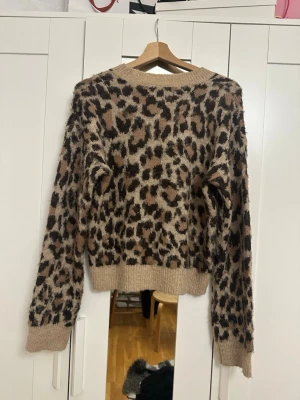 Leopardmönstrad stickad tröja - Säljer en fluffig stickad tröja med leopardmönster i beige, brunt och svart. Tröjan har långa ärmar, ribbade muddar och en avslappnad passform. Perfekt för dig som vill ha en trendig och mysig look.