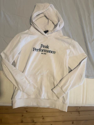 Vit hoodie från Peak Performance - Säljer en vit hoodie från Peak Performance med broderad logga och text 'Est. 1986' på bröstet. Hoodien har huva, känguruficka och ribbade muddar vid ärmslut och nederkant. Tillverkad i mjuk bomullsblandning, perfekt för en avslappnad stil.