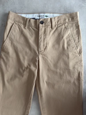 Beige chinos från Lacoste slim fit - Snygga beige chinos från Lacoste i slim fit-modell. Klassisk design med snedställda fickor fram och två bakfickor. Tillverkade i mjuk bomull med bälteshällor och knappgylf. Perfekta för en clean och stilren look.