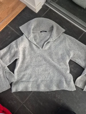 Ljusgrå stickad tröja med krage - Mysig ljusgrå stickad tröja med bred krage och v-ringning. Tröjan har vida ärmar och ribbstickade muddar vid ärmslut och nederkant. Perfekt för dig som gillar en avslappnad och trendig stil. Tröjan är lite nopprig.