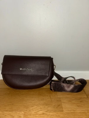 Mörkbrun axelväska - Valentino - BIGS - PRUGNA - Snygg mörkbrun axelväska från Valentino med justerbar axelrem och stilren design. Väskan har ett halvmåneformat lock med diskret logga i silver framtill. Tillverkad i slätt skinnliknande material som ger en lyxig känsla.