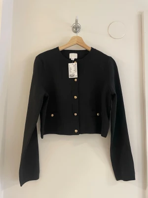 Svart kofta med guldfärgade knappar - Stilren svart kofta med rund halsringning och långa ärmar. 