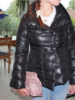 Svart pufferjacka med stora knappar - Snygg svart pufferjacka med stora dekorativa knappar framtill och bred krage. Jackan har quiltad design och en något figurnära passform. Perfekt för kalla dagar och ger en cool look till jeans eller andra outfits. Den är från BENETTON