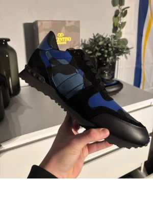 Valentino Rockrunner blå camo sneakers - Snygga Valentino Rockrunner sneakers i blått och svart med camouflagemönster. Skorna har svarta detaljer, snörning och platt sula. Tillverkade i en mix av skinn och syntetmaterial för en cool och modern look.