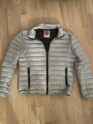 Vit/silvrig dunjacka från Colmar - Snygg dunjacka från Colmar med quiltad design och två svarta dragkedjeförsedda fickor framtill. Jackan har hög krage och hel dragkedja. Perfekt för kyliga dagar och enkel att matcha med olika outfits.