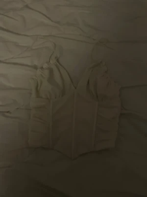 Vit bustier med rynkade axelband - Trendig vit bustier med rynkade axelband och markerad midja. Toppen har en figurnära passform och dekorativa sömmar framtill. Perfekt att styla under en skjorta eller som den är. Materialet känns mjukt och stretchigt.