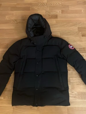 Svart dunjacka från Canada Goose - Snygg svart dunjacka från Canada Goose med huva och klassisk patch på ärmen. Jackan har en quiltad design, dragkedja och knappar framtill. Perfekt för kalla vinterdagar och riktigt varm. Passar dig som vill ha både stil och funktion.