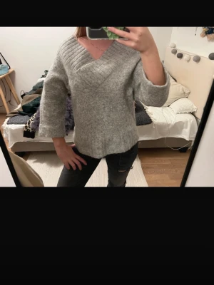 Grå stickad trekvartsärmad tröja med v-ringning - Mysig grå stickad tröja med bred v-ringning och ribbade detaljer vid hals, ärmslut och nederkant. Tröjan har en avslappnad passform och trekvartsärmar. Perfekt för lager på lager och chill dagar.