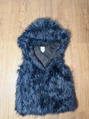 Mörkblå pälsväst med huva från GAP - Snygg mörkblå pälsväst från GAP Kids med fluffig fuskpäls och stor huva. Västen har ett mjukt innerfoder och är perfekt för att ge din outfit en cool touch. Passar dig som vill sticka ut och gillar trendiga detaljer.