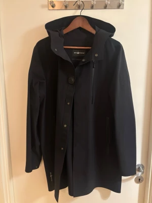 Sail Racing Regnkappa - Stilren svart regnjacka från Sail Racing med huva, dragkedja och knappstängning. Jackan har vattentäta fickor med dragkedja och diskret logga på ärmen. Perfekt för regniga dagar och enkel att matcha med olika outfits. Köpt från Sail Racing hemsida. Kvitto och förpackning förekommer. Nypris 3.500kr