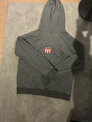 Grå hoodie från Gant med emblem - Mörkgrå hoodie från Gant med broderat emblem i rött, vitt och blått på bröstet. Klassisk huva och ribbade muddar vid ärmslut och nederkant. Perfekt för en avslappnad och sportig stil.