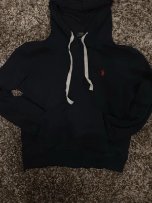 Mörkblå hoodie från Polo Ralph Lauren - Snygg mörkblå hoodie från Polo Ralph Lauren med vit dragsko och röd broderad logga på bröstet. Klassisk känguruficka framtill och huva. Tillverkad i mjukt bomullsmaterial för skön känsla. Perfekt för en avslappnad och stilren look.