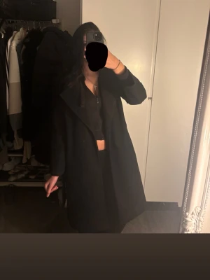 Svart kappa med klassisk krage - Säljer en svart kappa med klassisk krage och rak passform. Kappan har långa ärmar och en stilren look som passar perfekt till höst och vinter. Enkel design utan synliga detaljer som knappar. Perfekt för dig som gillar minimalistisk stil.