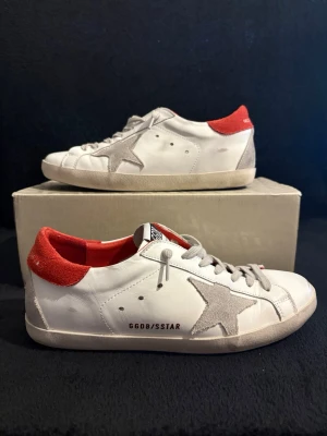 Golden Goose vita sneakers med röd häl - Golden Goose sneakers i vitt skinn med grå mockadetaljer och en stor stjärna på sidan. Röd mocka på hälen med logga, rund tå och snörning. Snygg kontrast mellan material och färger, perfekt för dig som gillar streetstyle.