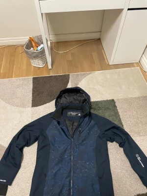 Mörkblå vindjacka från Ski Industries” pris kan diskuteras” - Snygg mörkblå vindjacka från Ski Industries med huva och hel dragkedja framtill. Jackan har diskret mönster på framsidan och är tillverkad i ett slitstarkt syntetmaterial. Perfekt för blåsiga dagar och outdoor-aktiviteter.