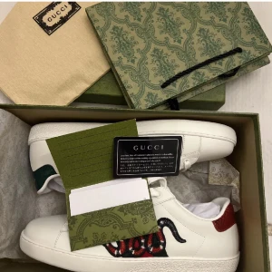 Gucci vita sneakers med orm brodyr - Säljer ett par vita Gucci sneakers i skinn med ikonisk orm brodyr i rött, svart och vitt på sidan. Skorna har grön och röd detalj på hälen, klassisk snörning och platt sula. Kommer med originalkartong och dustbag.
