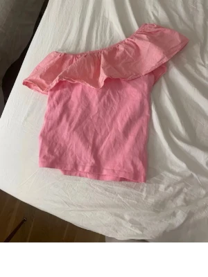 Rosa offshoulder topp kaphal - Snygg rosa offshoulder topp med stor volangdetalj längs överkanten. Perfekt för dig som vill sticka ut lite extra. Tillverkad i mjukt bomullstyg som känns skönt mot huden. Trendig och ungdomlig design. Storlek 158/162