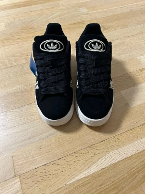 Adidas Campus svart/vit leopard - Snygga Adidas Campus sneakers i svart mocka med vita detaljer och leopardmönster på sidorna. Klassisk Adidas-logga på plösen och Campus-tryck på sidan. Tjock vit sula och svarta skosnören ger en clean look.