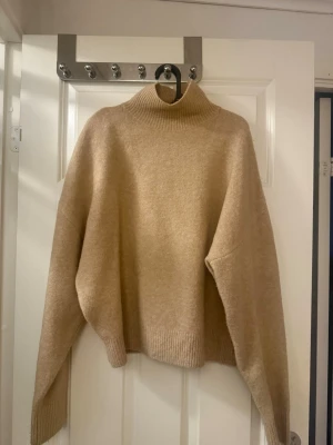 Beige stickad polotröja - Mysig beige stickad polotröja med avslappnad passform och ribbade muddar vid ärmslut, nederkant och hals. Tröjan har en hög polokrage och är tillverkad i ett mjukt material som passar perfekt för kyliga dagar.