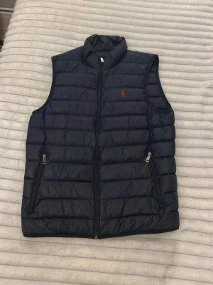 Mörkblå dunväst från Ralph Lauren - Snygg mörkblå dunväst från Ralph Lauren med diskret röd logga på bröstet. Västen har hög krage, två dragkedjeförsedda fickor och quiltad design. Perfekt att slänga över en hoodie eller tröja för extra värme och stil.