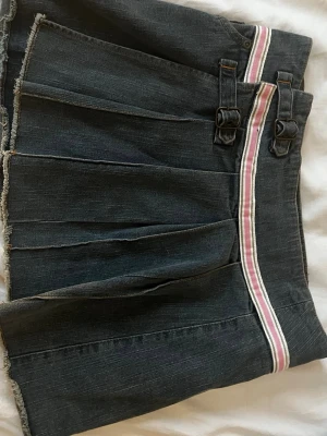Y2K, vintage Mörk jeanskjol med rosa rand - Säljer en mörkblå jeanskjol från Fishbone med rosa och vit rand i midjan. Kjolen har plisserade detaljer, två dekorativa spännen framtill och rå kant nertill för en cool look. Perfekt för dig som gillar streetstyle och denim.