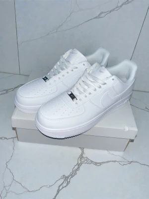 Nike Air Force 1 vita sneakers - Klassiska Nike Air Force 1 sneakers i helvitt läder med perforerad tå, vita skosnören och den ikoniska swoosh-loggan på sidan. Tjock sula med bra grepp och stilren design som passar till det mesta. Perfekta för dig som gillar en clean och tidlös look.