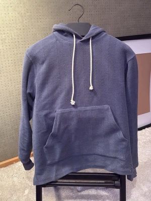 Blå hoodie från Aimé Leon Dore - Snygg blå hoodie från Aimé Leon Dore med vit dragsko och stor känguruficka framtill. Tillverkad i ett strukturerat tyg med diskret mönster. Perfekt för en avslappnad och trendig stil. Den har skön stretch och har en speciell designe lite över allt 