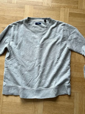 Grå sweatshirt från Gant - Säljer en stilren grå sweatshirt från Gant med broderad text 'Gant Girl' på bröstet. Tröjan har rund halsringning, ribbade muddar och är gjord i mjukt bomullsmaterial. Jätteskön.
