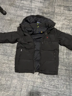 Svart pufferjacka från Polo Ralph Lauren - Säljer en svart pufferjacka från Polo Ralph Lauren med huva och dragkedja. Jackan har två fickor med dragkedja och den klassiska röda Polo-loggan på bröstet. Perfekt för kalla dagar och riktigt snygg till streetwear-stilen.
