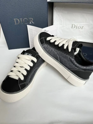 Dior grå och svarta sneakers - Snygga Dior sneakers i grått och svart med vit tjock sula och breda vita snören. Sidan har det klassiska Dior-mönstret och ovandelen är i en mix av textil och skinn. Perfekta för dig som vill ha en exklusiv och trendig look.