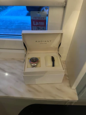 Radiant klocka i box med extra armband - Snygg klocka från Radiant i silverfärgat stål med svart urtavla och röd-blå vridring. Kommer i originalbox och med extra svart armband. Perfekt accessoar för dig som gillar stilrena detaljer och sportig look.