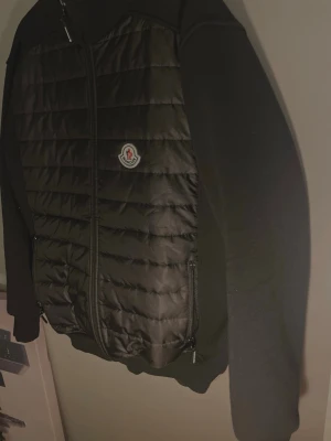 Svart cardigan jacka från Moncler - Svart cardigan jacka från Moncler med quiltad front och stickade ärmar. Jackan har dragkedja framtill, två sidofickor med dragkedja och Moncler-logga på bröstet. Perfekt för dig som vill ha en stilren och sportig look. Är lite för stor men skulle själv säga att det passar större än large och large lite vintage style och så