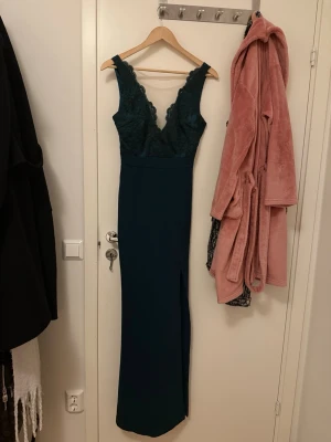 Mörkgrön långklänning med spets - Säljer en elegant mörkgrön långklänning med v-ringad spets upptill och breda axelband. Klänningen har en slits på sidan och figurnära passform, perfekt för speciella tillfällen. Materialet är troligtvis polyester med spetsdetaljer. Använd en gång och köpt ifrån Bubbleroom. Pris kan diskuteras!