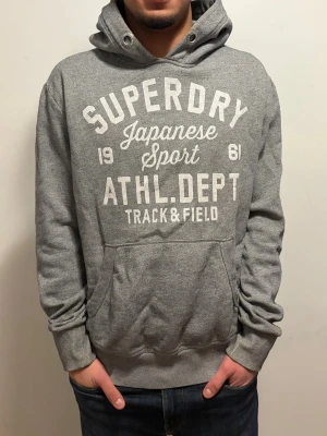 EXTREMT SÄLLSYNT Grå vintage Superdry hoodie - Extremt sällsynt Snygg grå hoodie från Superdry med stort vitt tryck på bröstet där det står 'Superdry Japanese Sport ATHL. DEPT Track & Field'. Klassisk känguruficka framtill och huva. Perfekt för en avslappnad och sportig stil.