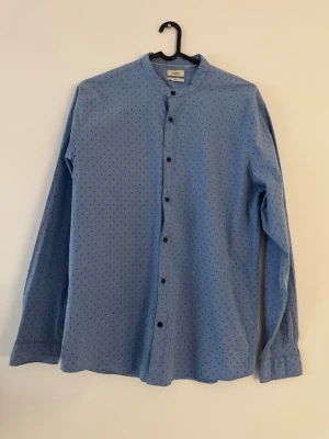 Blå prickig skjorta från Esprit - Snygg blå skjorta från Esprit med små svarta prickar och ståkrage. Skjortan har långa ärmar och svarta knappar framtill. Perfekt för dig som gillar en stilren men ändå unik look.