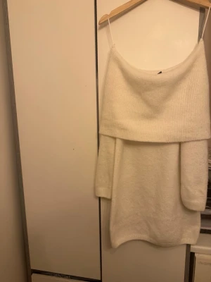 Vit offshoulder stickad klänning - Mysig vit stickad klänning med offshoulder-design och långa ärmar. Klänningen har en bred ribbad kant upptill och är perfekt för dig som gillar en avslappnad men ändå trendig stil. Passar bra till både sneakers och boots.