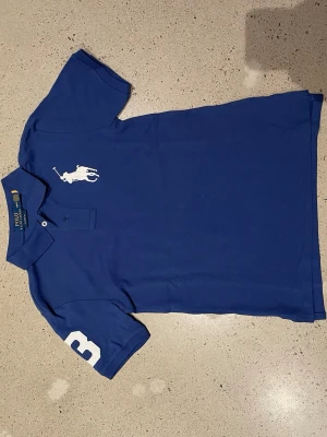 Blå pikétröja från Polo Ralph Lauren - Klassisk blå pikétröja från Polo Ralph Lauren med vit stor logga på bröstet och siffran 3 på ärmen. Tröjan har krage, knappar vid halsen och är tillverkad i bomull. Perfekt för en sportig och stilren look.