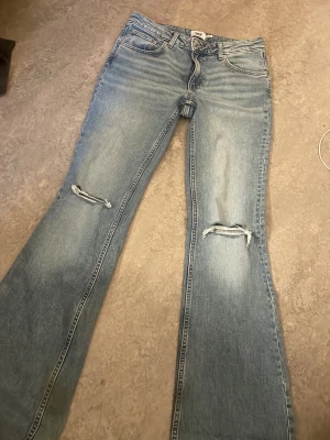 Blå bootcut jeans med slitningar - Snygga blå bootcut jeans med slitningar över knäna och klassisk femficksdesign. Jeansen har en ljus tvätt och raka ben som går ut något nedtill. Perfekta för en avslappnad och trendig look.